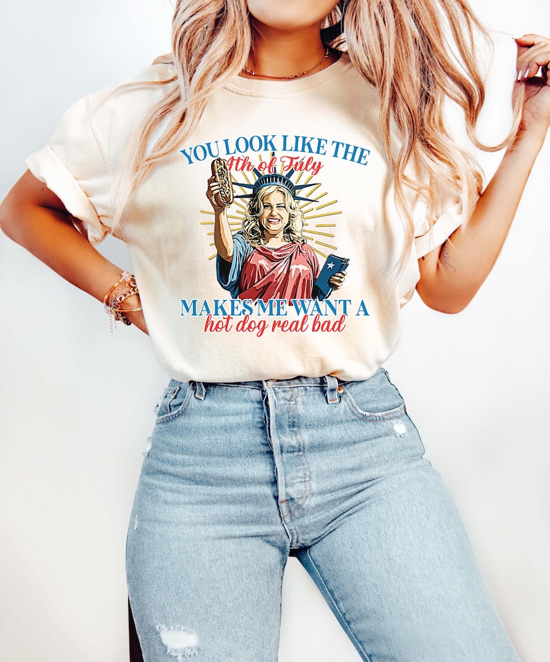 May include: Cream-colored t-shirt with a graphic of a woman dressed as the Statue of Liberty holding a hot dog. The text on the shirt reads "YOU LOOK LIKE THE 4th of July MAKES ME WANT A hot dog real bad."