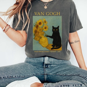 May include: Dark gray t-shirt featuring a print of Van Gogh's "Sunflowers (Third Version)" painting with a black cat. The text "VAN GOGH" is above the image, and "SUNFLOWERS (THIRD VERSION) - 1888" is below.