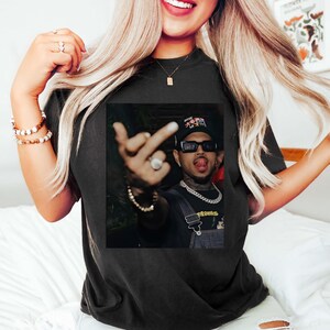 May include: Black t-shirt featuring a graphic of a person wearing sunglasses, a hat, and a chain. The person is making a hand gesture. The shirt is worn by a person with long blonde hair.