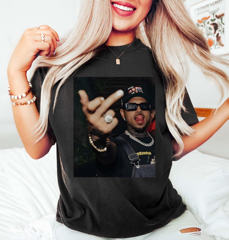 May include: Black t-shirt featuring a graphic of a person wearing sunglasses, a hat, and a chain. The person is making a hand gesture. The shirt is worn by a person with long blonde hair.
