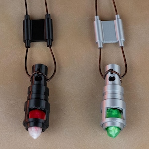 May include: Two pendant necklaces with brown cords. One is black with red and white accents, the other is silver with green accents. The pendants have a cylindrical shape. A silver and brown object is in the upper left corner.