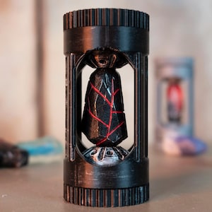 May include: A black, cylindrical decorative object with a geometric design. The object has a cage-like structure with a dark, angular centerpiece featuring red accents. The design is intricate and detailed, with a focus on sharp lines and contrasting colors.