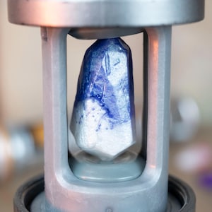 Peut inclure: Un cristal bleu et blanc à facettes est maintenu dans un cadre métallique argenté. Le cristal présente un effet de dégradé, passant du blanc à une teinte bleu profond. L'ensemble semble faire partie d'une machine ou d'un appareil.