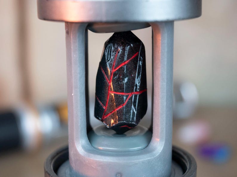 K&ouml;nnte beinhalten: Ein dunkler, facettierter Stein mit roten, venenartigen Mustern wird in einem silberfarbenen Metallrahmen gehalten. Der Stein ist der Mittelpunkt, wobei der Rahmen eine strukturierte Einfassung bietet. Der Hintergrund ist unscharf, was die Details hervorhebt.