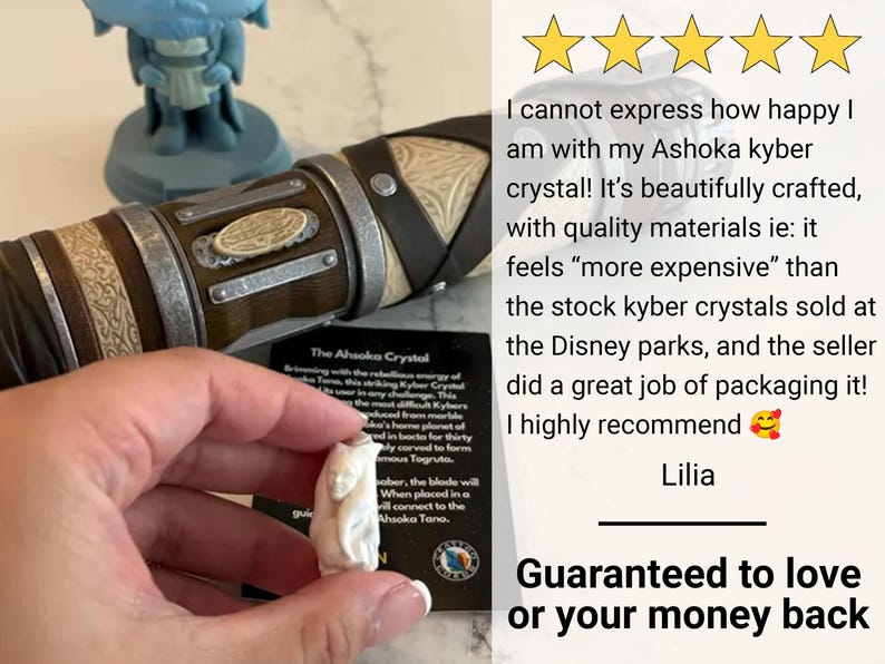 K&ouml;nnte beinhalten: Ein wei&szlig;er, geschnitzter Kristall, beschriftet mit "The Ahsoka Crystal", wird in der N&auml;he eines Lichtschwertgriffs gehalten. Das Bild enth&auml;lt eine Kundenrezension und eine Garantie.