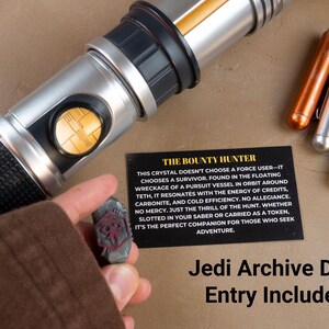 K&ouml;nnte beinhalten: Ein silbernes und schwarzes Lichtschwert mit goldenen Akzenten, begleitet von drei Metallstiften und einem kleinen, grauen Kristall. Eine schwarze Karte mit dem Text "The Bounty Hunter" ist ebenfalls vorhanden. Der Text "Jedi Archive Data Entry Included" befindet sich unten.