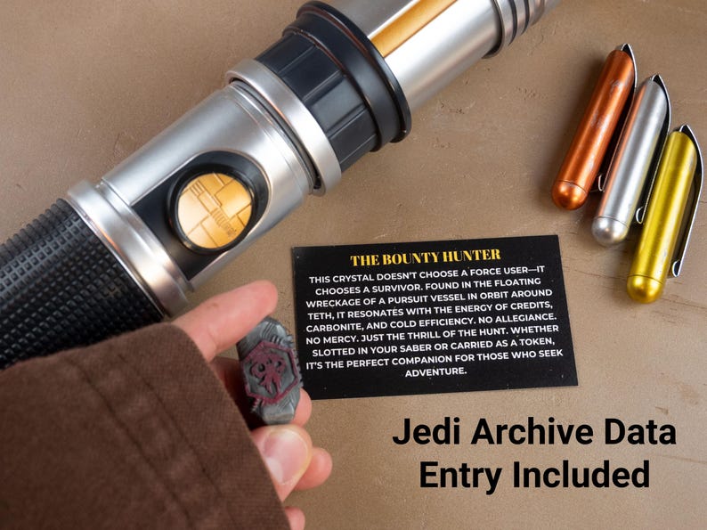 K&ouml;nnte beinhalten: Ein silbernes und schwarzes Lichtschwert mit goldenen Akzenten, begleitet von drei Metallstiften und einem kleinen, grauen Kristall. Eine schwarze Karte mit dem Text "The Bounty Hunter" ist ebenfalls vorhanden. Der Text "Jedi Archive Data Entry Included" befindet sich unten.