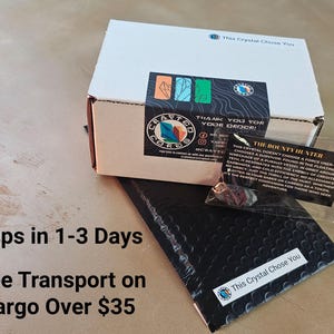 K&ouml;nnte beinhalten: Eine wei&szlig;e Schachtel mit einem schwarzen und farbenfrohen Aufkleber, einem schwarzen Luftpolsterumschlag und einem kleinen, durchsichtigen Beutel mit einer Karte. Der Text auf der Karte lautet "The Bounty Hunter". Auf der Schachtel und dem Umschlag steht der Text "This Crystal Chose You".