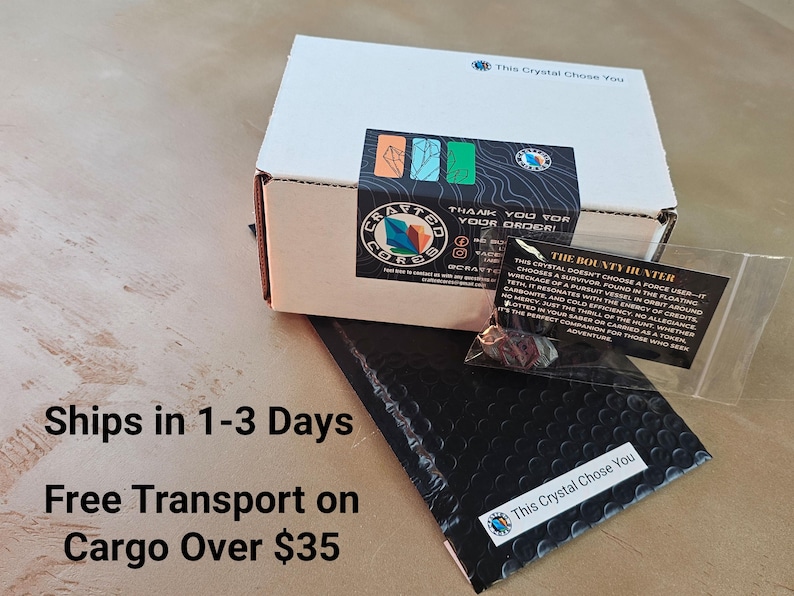 K&ouml;nnte beinhalten: Eine wei&szlig;e Schachtel mit einem schwarzen und farbenfrohen Aufkleber, einem schwarzen Luftpolsterumschlag und einem kleinen, durchsichtigen Beutel mit einer Karte. Der Text auf der Karte lautet "The Bounty Hunter". Auf der Schachtel und dem Umschlag steht der Text "This Crystal Chose You".