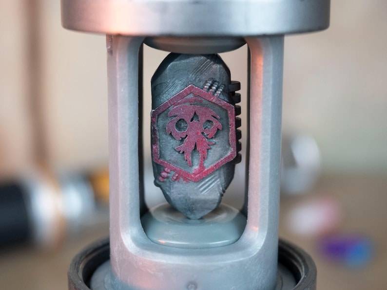 K&ouml;nnte beinhalten: Nahaufnahme eines metallisch grauen Objekts mit einem dunkelgrauen, steinartigen Mittelteil. Die Mitte zeigt ein rotes Emblem in einem sechseckigen Rahmen. Das Objekt ist in einen silbernen Rahmen eingefasst, was auf einen dekorativen oder funktionalen Gegenstand hindeutet.