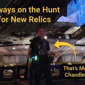 K&ouml;nnte beinhalten: Eine Person steht vor einer gro&szlig;en Raumschiffstruktur und h&auml;lt einen kleinen gelb-schwarzen Droiden. Das Bild enth&auml;lt den Text "Always on the Hunt for New Relics" und "That's Me! Chandler". Die Szene wird von blauen und gr&uuml;nen Lichtern beleuchtet.