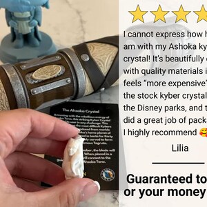 K&ouml;nnte beinhalten: Ein wei&szlig;er, geschnitzter Kristall, beschriftet mit "The Ahsoka Crystal", wird in der N&auml;he eines Lichtschwertgriffs gehalten. Das Bild enth&auml;lt eine Kundenrezension und eine Garantie.