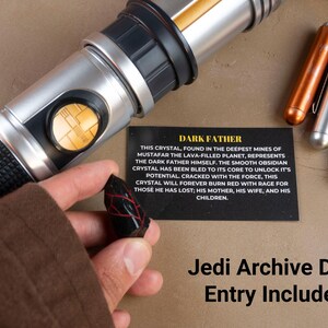 K&ouml;nnte beinhalten: Ein silbernes und schwarzes Lichtschwert mit einem goldenen Knopf, ein schwarzer Kristall mit roten Adern und drei metallische stiftartige Objekte in Kupfer, Silber und Gold. Eine Karte liest "DARK FATHER" und "Jedi Archive Data Entry Included."