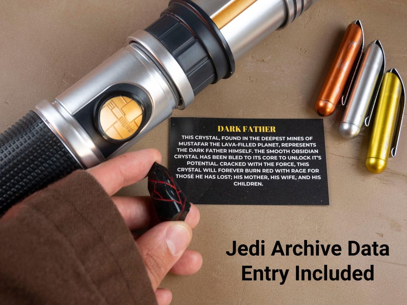 K&ouml;nnte beinhalten: Ein silbernes und schwarzes Lichtschwert mit einem goldenen Knopf, ein schwarzer Kristall mit roten Adern und drei metallische stiftartige Objekte in Kupfer, Silber und Gold. Eine Karte liest "DARK FATHER" und "Jedi Archive Data Entry Included."