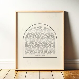May include: Framed print with a light wood finish. The artwork features a black text of a Bible verse from Matthew 5:2-12, arranged in a semi-circular shape. The background is a soft, off-white color.