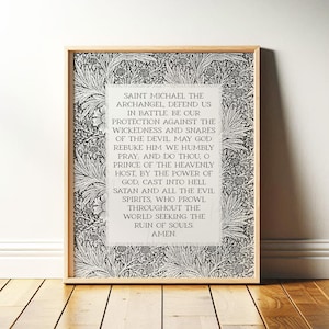 May include: Framed print featuring the text of a prayer to Saint Michael the Archangel. The text is centered on a white background, surrounded by a black and white floral pattern. The frame is a light wood color.