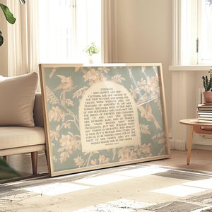 May include: A framed print with a floral design in pale blue and cream. The print has text within an arched shape, with a bird and floral pattern. The frame is light wood, and the print is leaning against a wall.