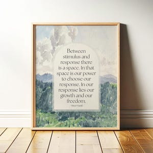 May include: Wooden framed art print with a watercolour landscape of mountains and clouds. The print features the quote, "Between stimulus and response there is a space..." by Viktor Frankl, in a white arched text box. The frame is light brown.