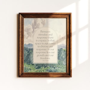May include: A framed print with a quote by Viktor Frankl. The artwork features a watercolor landscape with green hills and a cloudy sky. The quote reads, "Between stimulus and response there is a space..."