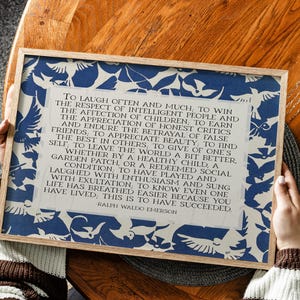 May include: A framed print featuring a quote by Ralph Waldo Emerson, surrounded by a blue and white bird pattern. The text is in a classic serif font. The frame is made of light-colored wood. The quote is about success.