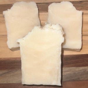 May include: Three rectangular bars of handmade soap with a creamy, off-white color. The edges of the soap have a slightly uneven, textured appearance. The soap bars are arranged on a wooden surface.