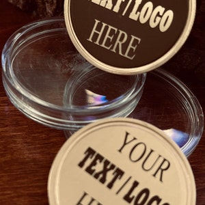 May include: Close-up of two custom coasters with the text "YOUR TEXT/LOGO HERE" in a bold font. One coaster is brown with a gold rim, and the other is off-white with a gold rim. Clear glass coasters are in the background.