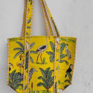Puede incluir: Bolso tote amarillo con un estampado de temática selvática con cebras, pájaros y palmeras. El bolso tiene asas amarillas a juego con detalles a rayas. El bolso está colgado contra una pared blanca.