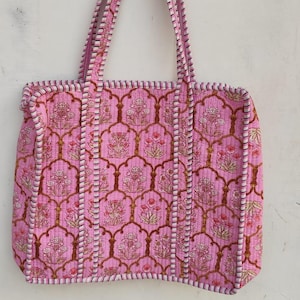 Puede incluir: Bolso tote acolchado rosa con un estampado floral. El bolso tiene forma rectangular con asas y ribetes a juego. El diseño floral presenta tonos rosas y marrones, creando un accesorio atractivo.