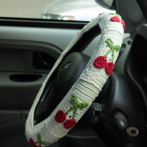 May include: A white crocheted steering wheel cover featuring a repeating pattern of small red cherries with green stems and leaves. The cover is fitted onto a black steering wheel inside a vehicle, showcasing a textured, handmade knit construction.