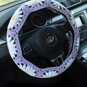 May include: A crochet steering wheel cover featuring a repeating pattern of white daisy motifs with yellow centers, connected by light purple yarn. The cover is fitted over a black steering wheel with a central silver Volkswagen logo in a vehicle interior.
