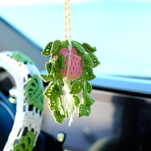 May include: A handmade crochet plant hanging from a car's rearview mirror. The plant pot is pink, surrounded by green leaves. The steering wheel has a white and green crochet cover. The background is a blurred view of the sky.