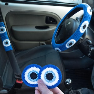 May include: A set of blue and white crocheted evil eye charms for a car. The charms are in the shape of a circle with a blue eye in the center. The charms are attached to the steering wheel, gear shift, and door handle.