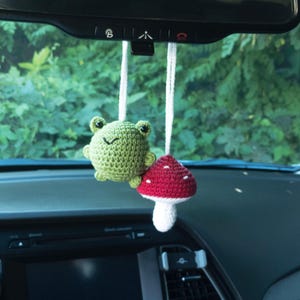 May include: A green crocheted frog and a red crocheted mushroom hanging from a rearview mirror in a car.