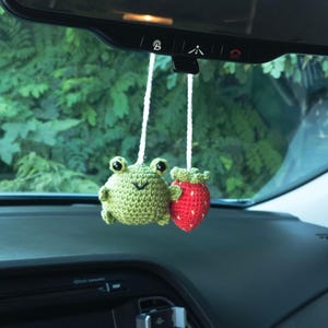May include: A green crocheted frog and a red crocheted strawberry hanging from a rearview mirror in a car.