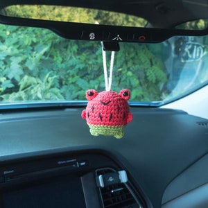 May include: A crocheted watermelon frog hanging from a rearview mirror in a car. The frog is red and green and has a white string attached to it.