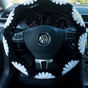 K&ouml;nnte beinhalten: Schwarze Lenkradabdeckung mit wei&szlig;en G&auml;nsebl&uuml;mchen. Die Abdeckung ist geh&auml;kelt und hat ein schwarz-wei&szlig;es Muster. Das Lenkrad hat eine schwarze Lederbezug und ein silbernes Airbag-Etikett.