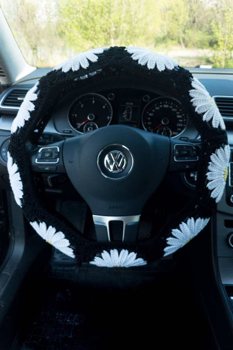 K&ouml;nnte beinhalten: Schwarze Lenkradabdeckung mit wei&szlig;en G&auml;nsebl&uuml;mchen. Die Abdeckung ist geh&auml;kelt und hat ein schwarz-wei&szlig;es Muster. Das Lenkrad hat eine schwarze Lederbezug und ein silbernes Airbag-Etikett.