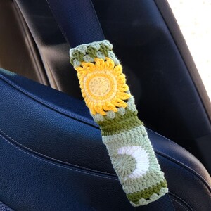 May include: Crocheted seatbelt cover featuring a yellow sunflower design and a white crescent moon. The cover is made with green, yellow, and white yarn and is designed to fit over a car seatbelt. The background shows a black car seat.