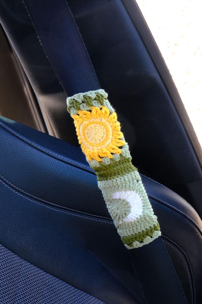 May include: Crocheted seatbelt cover featuring a yellow sunflower design and a white crescent moon. The cover is made with green, yellow, and white yarn and is designed to fit over a car seatbelt. The background shows a black car seat.