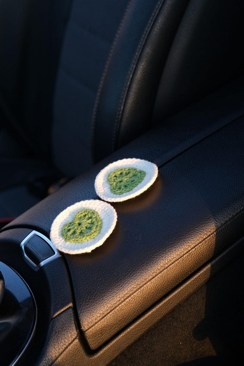 Pair of white and green crochet car coasters with Monstera leaf design on a car center console.