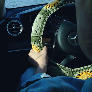 May include: A green and yellow crocheted steering wheel cover with a sunflower design. The cover is on a black steering wheel in a car, with a view of the ocean in the background.