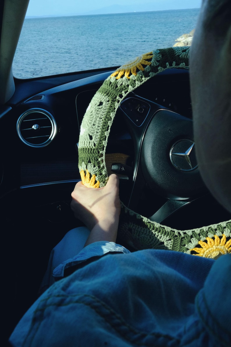 May include: A green and yellow crocheted steering wheel cover with a sunflower design. The cover is on a black steering wheel in a car, with a view of the ocean in the background.