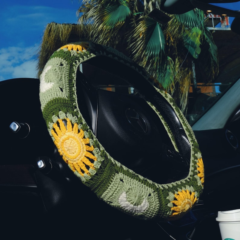 May include: A crocheted steering wheel cover featuring a sunflower design with yellow and green hues. The cover is placed on a black steering wheel inside a car, with a palm tree visible in the background.