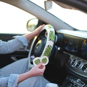 Monstera crochet steering wheel cover in a car while matching green and white crochet coasters are held in hand.