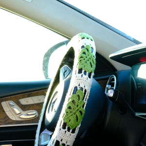 White and green Monstera crochet steering wheel cover fitted on a car wheel in bright natural light.