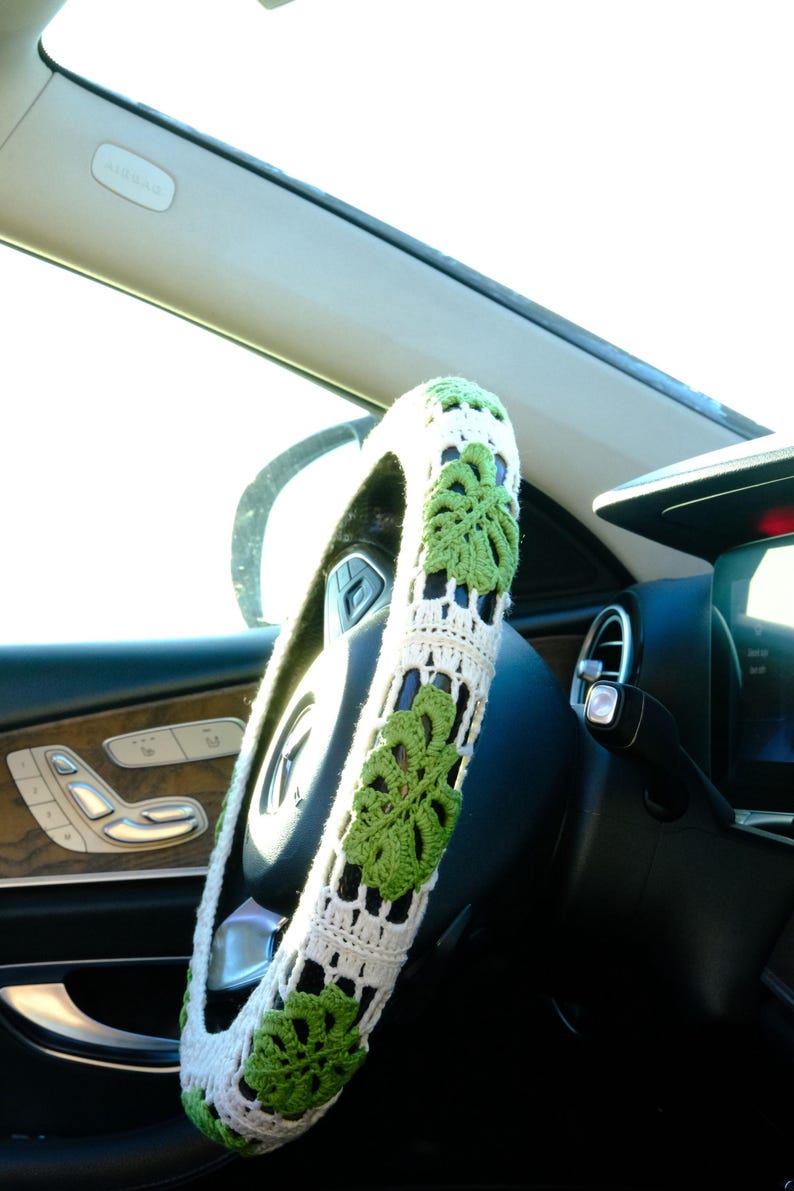 White and green Monstera crochet steering wheel cover fitted on a car wheel in bright natural light.