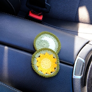 May include: Two handmade crochet coasters. One features a yellow sunflower design with a green border, and the other has a white swirl on a green background. These decorative coasters are perfect for protecting surfaces from drink condensation.