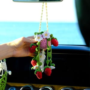 May include: A handmade crochet car charm featuring a pink pot with white flowers and red strawberries. The charm is suspended by a cream-colored string. The charm is held in front of a car dashboard and a blue ocean background.