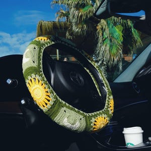 May include: Crocheted steering wheel cover featuring a sunflower design. The cover is made of green, yellow, and white yarn, adding a touch of handmade charm to the car's interior. The background includes a palm tree and a blue sky.