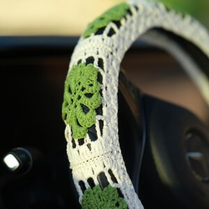 Close-up of a white crochet steering wheel cover decorated with textured green Monstera leaves.